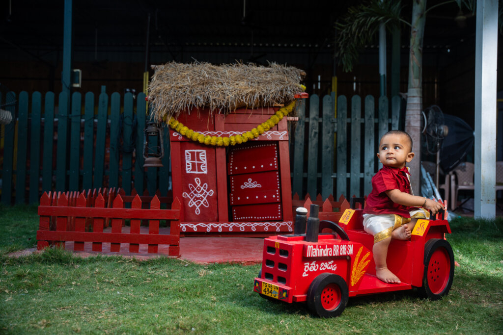 A young boy dressed as a farmer, sitting in a toy tractor surrounded by straw, hay bales, and festive farm-themed decorations for his pre-birthday shoot.