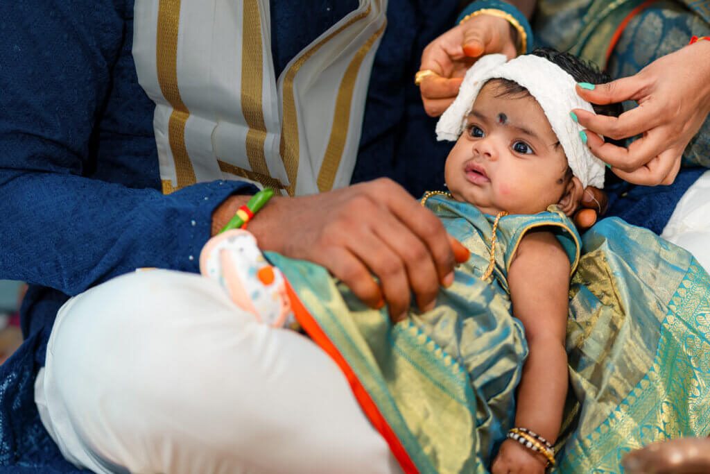 Baby girl sitting peacefully in her father's lap during the cradle ceremony, both surrounded by warm light and traditional decor.