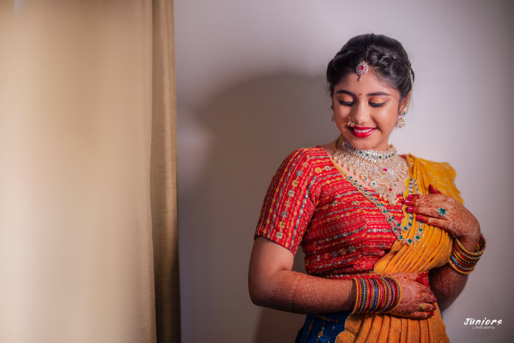 Wide portrait of a young girl in a bright saree during her saree ceremony, smiling gracefully, with traditional decor and family members blurred in the background.
